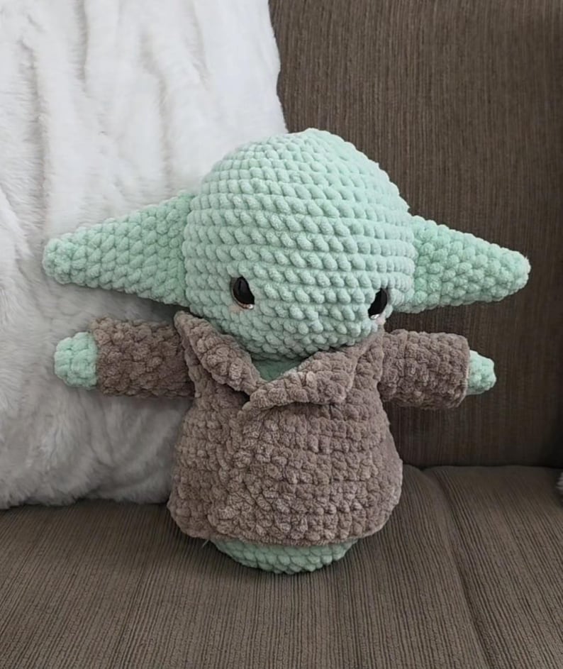 May include: A crocheted plush toy in the likeness of a character with large ears. The toy is primarily light green with a brown robe. The toy has black eyes and is sitting on a brown surface.