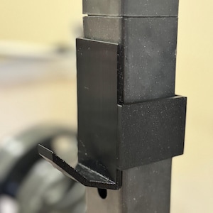 May include: Black weightlifting rack attachment. The attachment is designed to hold a barbell. The attachment is made of black metal and has a hook shape. The rack has several holes for adjustment.
