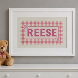 May include: A framed cross-stitch embroidery featuring the name REESE in dark pink capital letters, centered within a pink and white gingham checkered border with small bow accents at the top left and bottom right corners, set in a white frame.