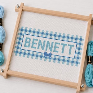 May include: A square wooden embroidery frame holding white fabric with a cross-stitch design of the name BENNETT in blue letters, surrounded by a blue and white checkered border and a small blue bow, flanked by several skeins of blue embroidery floss.
