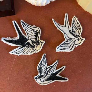 Könnte beinhalten: Drei gestickte Aufnäher mit einem Schwalben-Design in Schwarz und Weiß. Die Aufnäher zeigen Schwalben im Flug. Die Aufnäher liegen auf einer rotbraunen Oberfläche.