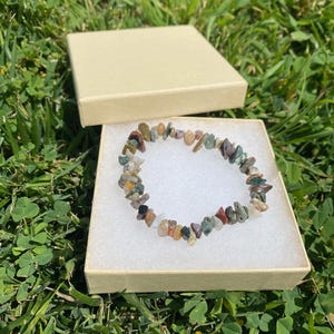 May include: A multi-colored stone chip bracelet rests inside a beige gift box with a white interior. The bracelet features a variety of colors, including green, brown, red, and white. The box is open, and the bracelet is displayed on a bed of white material.