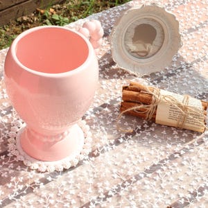 Puede incluir: Una taza de cerámica rosa con un asa decorativa se encuentra sobre un tapete de encaje blanco. Una pequeña silueta enmarcada en blanco y un manojo de palitos de canela atados con hilo también están sobre la mesa.