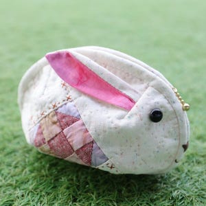 Könnte beinhalten: Ein handgefertigter, hasenförmiger Beutel aus gestepptem Stoff. Der Beutel hat einen cremefarbenen Körper mit einem rosa Innenohr und einem Patchwork-Design an der Seite. Er hat ein schwarzes Knopfauge und eine kleine goldene Kette.