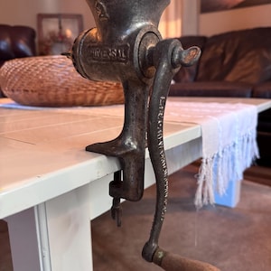 May include: An antique Universal meat grinder made of cast iron. The grinder is a dark, aged metal with a wooden handle. The number "2" is visible on the top. The grinder is clamped to a white table.
