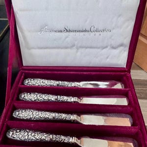 May include: A set of four butter knives in a burgundy velvet box. The knives have ornate floral handles and shiny silver blades. The box is lined with white satin and features the text "American Silversmiths Collection".