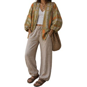 May include: A woman's outfit featuring a patterned kimono in shades of green, orange, and beige, paired with a white top and beige linen pants. The outfit is completed with brown sandals and a woven bag.