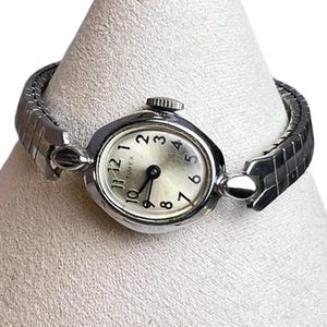 May include: A vintage Timex watch with a silver-toned face and a silver expanding band. The watch face displays black numerals and hands against a light background. The watch is displayed on a white surface.