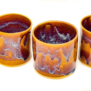 May include: Three handmade ceramic cups with a cylindrical shape. The cups have a glossy finish with a blend of orange, brown, and purple hues. The glaze drips down the sides, creating a unique texture. The cups are set against a white background.