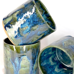 May include: A stack of four handmade ceramic tumblers. The exterior features a blend of blue and green glaze with a dripped effect. The interior of the cups is a deep blue. The cups are cylindrical with a slightly flared rim.