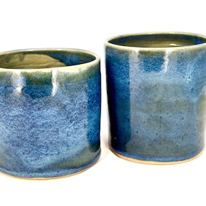 Puede incluir: Dos vasos de cerámica hechos a mano con un esmalte azul y verde. Las tazas cilíndricas tienen una superficie texturizada y un borde y base de color claro. Las tazas miden aproximadamente 10 cm de alto y 8 cm de diámetro.