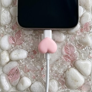 Può includere: Un organizzatore di cavi a forma di cuore rosa è attaccato a un cavo di ricarica bianco, collegato a uno smartphone nero. Il telefono poggia su una superficie decorativa con pietre bianche, cristalli trasparenti e accenti di foglie rosa.