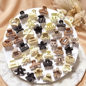 Könnte beinhalten: Eine Sammlung kleiner Haarspangen in verschiedenen Formen und Farben, darunter Blumen, Sterne und Herzen. Die Spangen sind in Schwarz-, Creme- und Brauntönen auf einer weißen, kreisförmigen Oberfläche angeordnet.