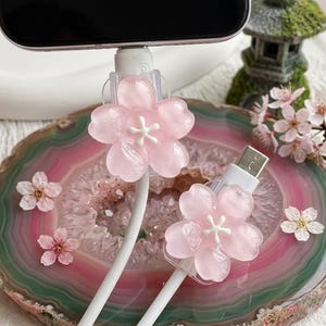 May include: Two white charging cables with pink cherry blossom cable protectors. The blossoms have a white centre and are set against a pink and green agate slice. Scattered cherry blossoms add to the floral theme.