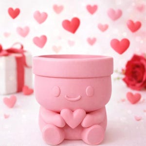 Könnte beinhalten: Ein hellrosa Blumentopf in Form einer Zeichentrickfigur, die ein Herz hält. Der Topf hat ein lächelndes Gesicht und ist von schwebenden Herzformen umgeben. Eine Geschenkbox und eine Rose befinden sich im Hintergrund.