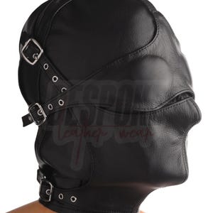 Puede incluir: Capucha de cuero negro con múltiples correas y hebillas metálicas. La capucha cubre toda la cabeza y el cuello, con aberturas para los ojos y la boca. La capucha está diseñada con una forma contorneada y se asegura con correas ajustables.