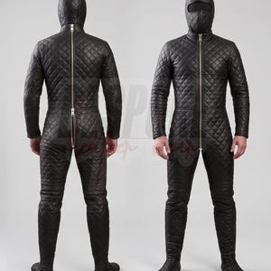 Puede incluir: Un traje de cuerpo entero de cuero negro acolchado con capucha, abertura facial y una larga cremallera plateada en la parte delantera y trasera. La prenda cubre los brazos, el torso, las piernas y los pies con un patr&oacute;n de costuras.