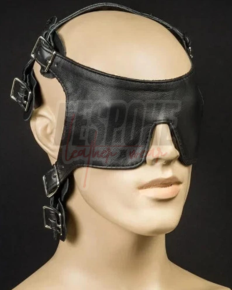 Puede incluir: Antifaz de cuero negro con correas y hebillas ajustables. El antifaz cubre los ojos y tiene una correa que rodea la cabeza. El texto "BESPOKE Leather" es visible en el antifaz.