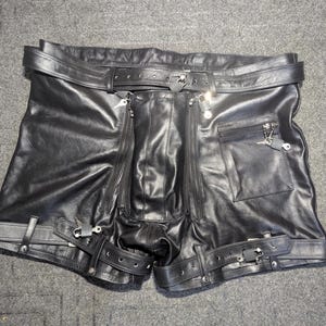 Op de afbeelding: Zwarte leren shorts met een tailleband met gesp, twee verticale ritszakken aan de voorzijde en een vierkante ritszak aan de rechterkant. De beenopeningen zijn versierd met verstelbare gespriemen en metalen hardware-accenten.