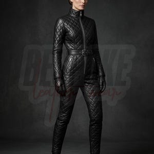 Puede incluir: Un mono de cuero negro acolchado con cuello alto, cremallera frontal completa y cinturón a juego con hebilla. La prenda presenta mangas largas, pantalones ajustados y un patrón de costuras en forma de diamante en toda la pieza.