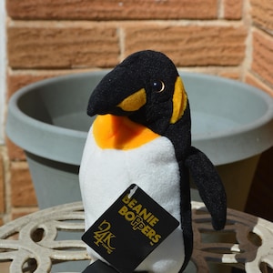 Könnte beinhalten: Ein Plüschpinguin mit schwarzem Kopf, Flügeln und Füßen, einem weißen Körper und gelben Akzenten. Der Pinguin hat ein Etikett mit der Aufschrift "Beanie Boppers" und "24K Company". Das Spielzeug steht auf einem weißen Metalltisch.