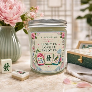 May include: A clear glass candle jar with a silver lid, featuring a label that reads A SCENTING CANDLE COMPANY, LIGHT IT. LOVE IT. ENJOY IT. The label displays mahjong tiles, pink flowers, and bamboo. It is marked 9 OZ | 255 G.