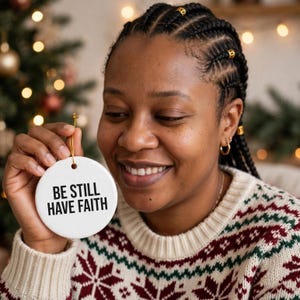 Op de afbeelding: Een rond wit keramieken ornament met de zwarte tekst BE STILL HAVE FAITH. Het ornament hangt aan een dun gouden koordje en heeft een gladde, glanzende afwerking, ontworpen om in een boom te hangen of als decoratief accentstuk.