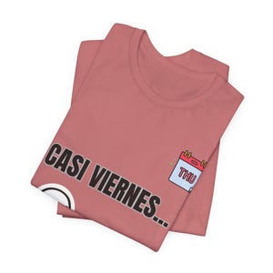 May include: A dusty rose t-shirt featuring the black text CASI VIERNES... and a small graphic of a calendar page showing the letters THU in red. The shirt is folded, displaying a crew neckline and short sleeves on a plain background.