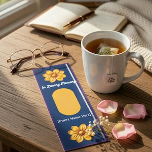 Puede incluir: Un marcapáginas azul marino con diseños florales amarillos y el texto "In Loving Memory" y espacio para un nombre. Una taza de té blanca, gafas y un libro abierto están sobre una mesa de madera.