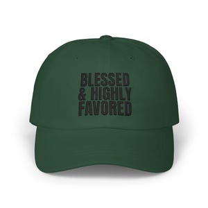 May include: A dark forest green baseball cap featuring the embroidered black text BLESSED & HIGHLY FAVORED centred on the front panel. The cap has a structured crown, a curved brim, and a button at the top, presenting a clean and simple design.