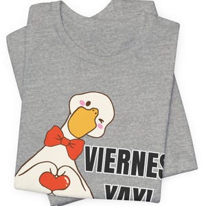May include: A heather grey t-shirt featuring a cartoon goose wearing a red bow tie. The goose is making a heart shape with its wings and holding a red heart. The text VIERNES YAY is printed in bold black letters with a white outline on the right side.
