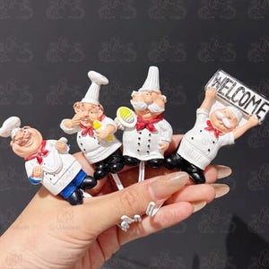 Può includere: Quattro statuette decorative di chef, ognuna con un cappello e una divisa bianchi con un fazzoletto rosso. Uno chef tiene in mano un utensile giallo, un altro un piatto e il terzo tiene un cartello con la parola WELCOME. Base a gancio bianca.