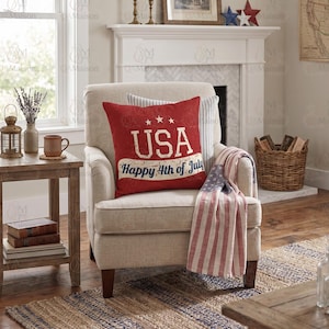 May include: A square red throw cushion featuring the white text USA and Happy 4th of July with three stars above the lettering. The cushion is placed on a beige upholstered armchair alongside a folded throw blanket with a red, white, and blue flag pattern.