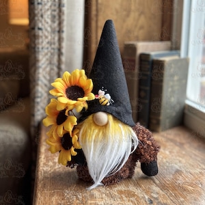 May include: A gnome figurine featuring a tall black felt hat adorned with a small bee embellishment. The gnome has a round wooden nose, a long white beard, and a textured brown body. A cluster of three yellow sunflowers is attached to the side.