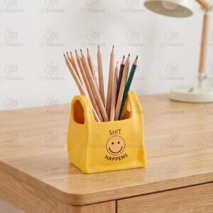 May include: A yellow ceramic pencil holder shaped like a small shopping bag, featuring a black printed smiley face and the text "SHIT HAPPENS" on the front. The holder is filled with a collection of wooden pencils in various natural and colored tones.