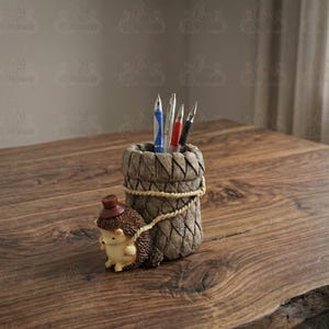 Puede incluir: Un portalápices decorativo hecho de material tejido, con un detalle de cuerda y una pequeña figura de erizo. El portalápices contiene varios bolígrafos y lápices. El erizo lleva un sombrero y parece estar tirando del portalápices.