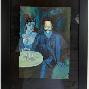Puede incluir: Una impresión enmarcada de una pintura que representa a un hombre y una mujer sentados a una mesa con una jarra y un vaso. Las figuras están representadas en tonos azules y verdes. El hombre tiene bigote y la firma Picasso es visible.