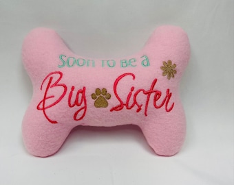 May include: A light pink, bone-shaped plush toy featuring embroidered text that reads Soon to be a Big Sister in teal and red thread. The design includes a small brown paw print icon between the words and a brown flower icon on the right side.