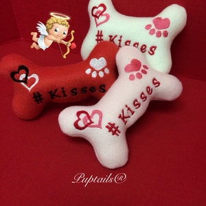 Könnte beinhalten: Drei Plüsch-Hundespielzeug in Form von Knochen in Rot, Weiß und Rosa mit dem aufgenähten Text "# Kisses". Die Spielzeuge haben ein Herz- und Pfotenmuster.