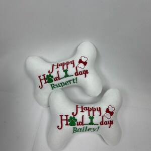 May include: Two white plush dog bone toys with red and green embroidery. The text on the toys reads "Happy Howl-idays" with a paw print and bone graphic. The names "Rupert!" and "Bailey!" are embroidered on the toys.