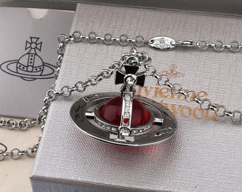 May include: A silver-toned necklace featuring a pendant shaped like a Saturn-inspired orb. The orb has a deep red translucent center, a silver ring encircling it, and a cross topper adorned with small clear crystals and a red accent stone.