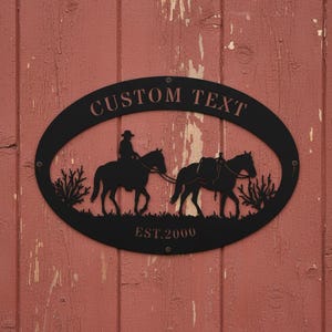 Könnte beinhalten: Schwarze Metall-Wandkunst mit einem Cowboy auf einem Pferd und einem Packpferd, mit dem Text "CUSTOM TEXT" und "EST.2000" in einem ovalen Rahmen. Auf einer verwitterten roten Holzoberfläche montiert.
