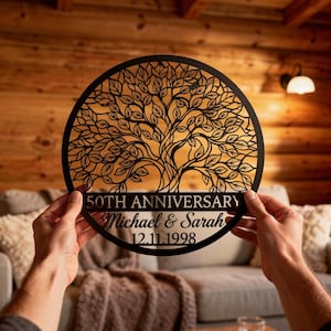 Könnte beinhalten: Ein kreisförmiges Wandkunstobjekt aus schwarzem Metall mit einem detaillierten, lasergeschnittenen Baummotiv. Der Text 50TH ANNIVERSARY, Michael & Sarah und 12.11.1998 ist im unteren Teil des Kreises eingraviert, was eine personalisierte Gedenktafel ergibt.