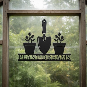 Könnte beinhalten: Schwarzes Metall-Gartenkunstwerk mit einer Kelle, zwei Blumentöpfen und dem Schriftzug "PLANT DREAMS". Das Kunstwerk ist vor einem verschwommenen grünen Hintergrund platziert, der einen Garten andeutet. Das Design ist schlicht und elegant.