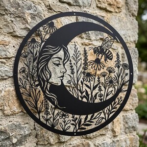 Könnte beinhalten: Ein kreisförmiges Wandkunstobjekt aus schwarzem Metall mit einem ausgeschnittenen Design aus einer Mondsichel, die in ein Gesichtsprofil integriert ist, umgeben von verschiedenen Wildblumen und einer Biene. Die Silhouette ist aus Metall gefertigt.