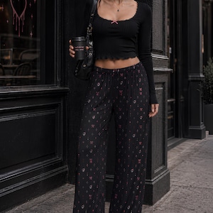 Può includere: Top nero a maniche lunghe con dettaglio fiocco rosa e pantaloni neri a gamba larga con motivo a fiocchi rosa. L'outfit è abbinato a scarpe nere e una borsa a tracolla nera. Viene tenuta una tazza di caffè.