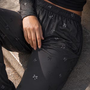 May include: Black pajama pants with a silver bow pattern. The pants have an elastic waistband and are paired with a black long-sleeve top. The fabric appears to be soft and flowing, suitable for sleepwear or loungewear.