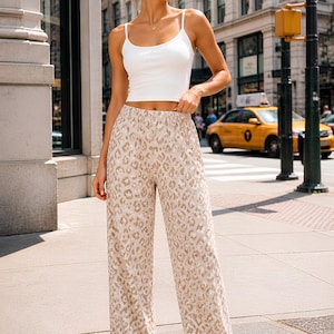May include: A woman wearing a white spaghetti strap top and wide-leg pants with a tan and brown animal print. She is wearing white sneakers. The background includes a city street with buildings and a yellow taxi cab.