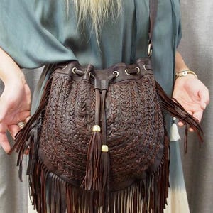 Op de afbeelding: Een bruine leren schoudertas met een geweven ontwerp en franjes. De tas heeft een trekkoordsluiting en decoratieve kwastjes. De tas wordt gedragen met een groene top en een crèmekleurige rok.