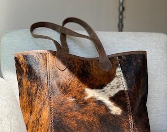 Puede incluir: Un bolso tote grande con un estampado de piel de vaca en tonos marrones, negros y blancos. El bolso tiene dos asas de cuero marrón y un ribete de cuero marrón. El bolso está sobre una silla gris tapizada.